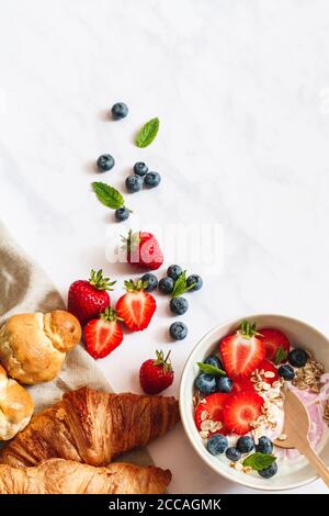 Petit déjeuner en été. Bol de yaourt avec fraises, bleuets et muesli, croissants et brioche parisienne sur fond de marbre blanc. Banque D'Images