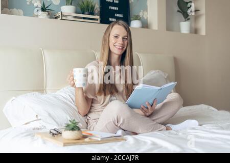 Heureuse et souriante détendue gaie femme lisant le carnet, travaillant à la maison bureau, s'assoit sur le lit et boire du café. Blonde fille appréciant gratuitement Banque D'Images