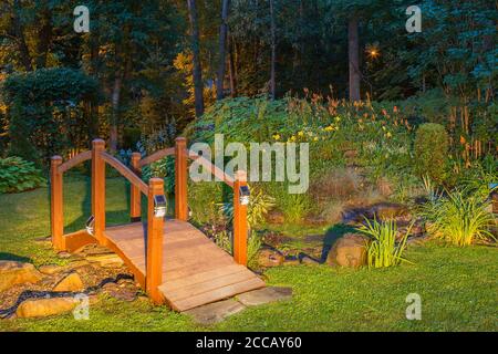 Passerelle au-dessus du ruisseau et de l'étang au crépuscule bordée de plantes et d'arbustes vivaces, y compris Hemérocallis orange et jaune - fleurs de Daylis, Hosta. Banque D'Images