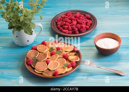 Céréales pancakes et framboises fraîches mûres rouges dans une assiette brune. Un bouquet de Melissa dans une tasse sur une table en bois bleu. Délicieux petit déjeuner d'été Banque D'Images