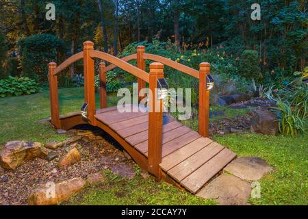 Passerelle au-dessus du ruisseau et de l'étang au crépuscule bordée de plantes et d'arbustes vivaces, y compris Hemérocallis orange et jaune - fleurs de Daylis, Hosta. Banque D'Images