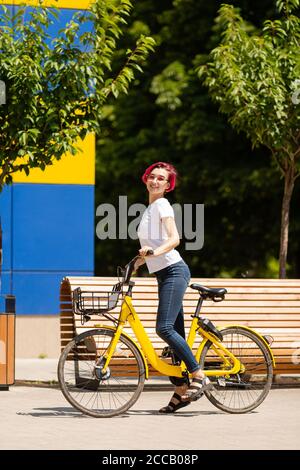 Une jeune femme heureuse avec des cheveux roses marche dans la ville sur un vélo en été. Mode de transport écologique Banque D'Images