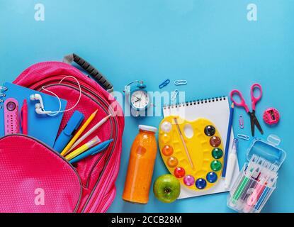Sac à dos rose, bouteille de smoothie aux fruits et papeterie sur fond bleu. Vue de dessus, espace de copie. Banque D'Images