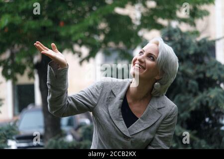 Bonne femme blonde mûre marche dans la pluie et attrape des gouttes avec la main, foyer sélectif Banque D'Images