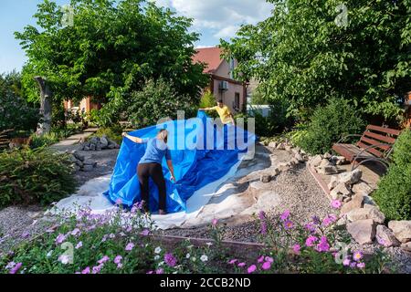 La famille pose un film plastique HDPE bleu sur le sol pour installer un étang à poissons Banque D'Images