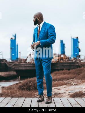 Un homme noir à barbe chauve à la mode, debout à l'extérieur dans un costume bleu élégant avec une cravate, dans des chaussures en cuir, avec trois constructions bleues dans Banque D'Images