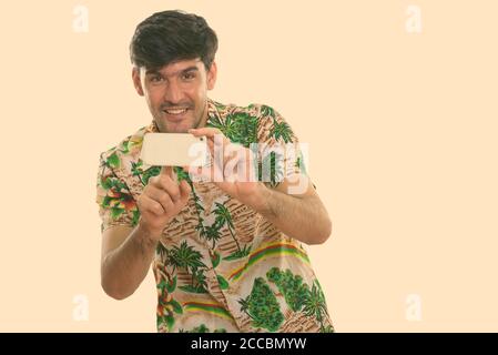 Studio shot of young happy man smiling persique pendant la prise de photo avec un téléphone mobile Banque D'Images