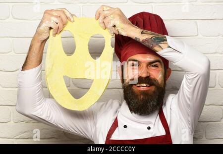 Homme avec barbe tient la pâte roulée avec smiley coupé sur fond de mur de brique blanche. Faites cuire avec un visage excités dans des pièces d'uniforme bordeaux avec de la pâte à pizza. Le chef prépare de la pâte. Concept de cuisine professionnelle. Banque D'Images
