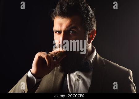 Homme avec barbe tient le cigare sur fond noir. Guy en costume classique a cinq minutes de pause pour fumer. Un homme d'affaires au visage sérieux fume un cigare cubain. Concept d'affaires et de mauvaise habitude. Banque D'Images