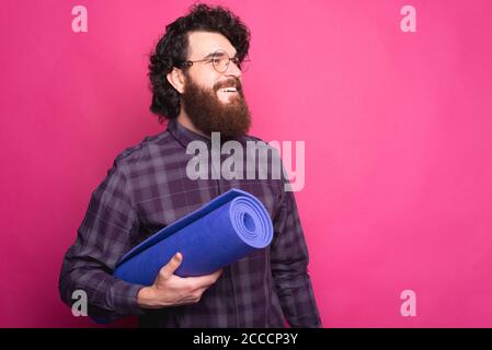 Photo de l'homme hipster barbu souriant tenant un tapis de yoga violet et regardant loin. Banque D'Images