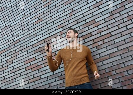 Portrait d'un homme souriant avec un casque et un téléphone portable debout près d'un mur de briques. Banque D'Images