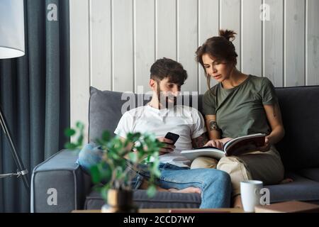 Un jeune couple charmant se détendant sur un canapé à la maison, lisant un magazine, utilisant un téléphone portable Banque D'Images