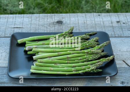 Bouquet d'asperges vertes fraîches dans une assiette noire sur une table rustique en bois été à l'extérieur. Banque D'Images