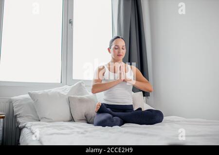 consciente femme caucasienne dans lotus poser sur le lit le matin, méditant femme assis avec les yeux fermés, isolé dans la chambre à coucher lumineuse Banque D'Images