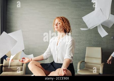 Calme jeune femme aux cheveux rouges vêtue en tenue formelle ayant la méditation de yoga sur la table au bureau étant entourée de papiers volants. Faire une pause pour se détendre Banque D'Images