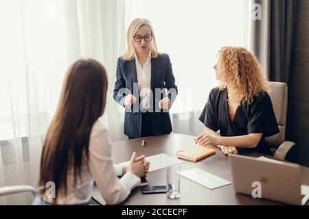 Les femmes d'affaires habillées avec élégance au bureau ayant des conversations et utilisant la technologie, se concentrent sur la femme blonde en train de dire quelque chose à ses collègues. Banque D'Images