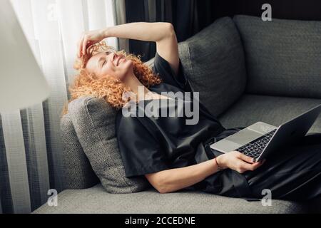 Busness femme avec des cheveux rouges de curly ayant rêvant l'expression heureuse tout en vous relaxant avec un ordinateur portable sur le canapé de bureau pendant la pause déjeuner Banque D'Images