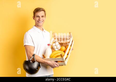 portrait de jeune coursier souriant transportant des aliments frais, des légumes et des fruits, portant un uniforme blanc avec un casque pour moto, concep de livraison express Banque D'Images