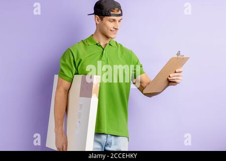 portrait d'un jeune homme de livraison caucasien souriant en chemise verte, d'un beau pizaman tenant une boîte avec pizza et regardant la liste des ensembles de pizza commandés. c Banque D'Images