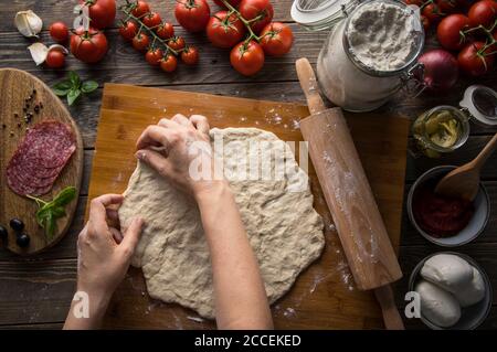 Une femme préparant une pizza, pétrissez la pâte et mettez les ingrédients sur une table en bois de cuisine. Sombre et moody, prise de vue aérienne. Banque D'Images