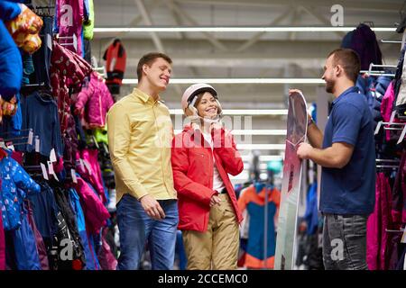 sourire couple caucasien écouter un agréable vendeur parler de snowboard, jeune femme veulent acheter un casque et d'autres équipements pour le ski, regardez s Banque D'Images