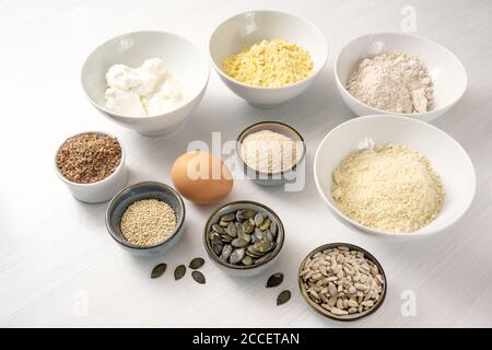 Ingrédients pour un pain aux protéines avec du fromage blanc, du son d'avoine, de la farine de lupin, de l'amande et de diverses graines dans des bols sur une table blanche, cuisson saine pour le carb bas ou Banque D'Images