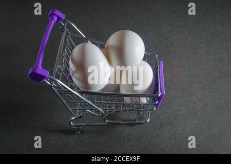 Chariot d'épicerie avec trois œufs de poulet blancs sur fond gris foncé. Banque D'Images