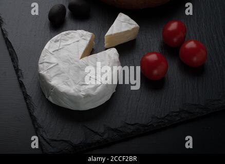 Le Brie frais et une tranche sur une assiette en pierre noire avec tomates cerises, olives et pain sur la table noire Banque D'Images