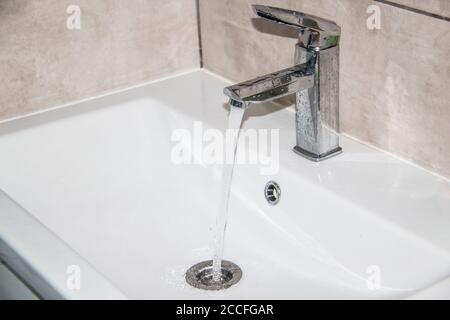 De l'eau coule du robinet vers l'évier. Robinet d'eau moderne dans la salle de bains avec eau courante dans le lavabo. Ouvrez le robinet de l'évier chromé. Chauve-souris au design moderne Banque D'Images