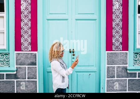 Belle femme aux cheveux blonds bouclés en utilisant le téléphone intelligent dans la rue - porte bleu clair en arrière-plan Banque D'Images