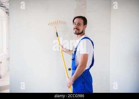 Un homme barbu en combinaison de travail peint le mur avec un rouleau. Il sourit amical et pose pour la caméra Banque D'Images