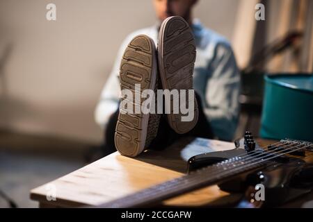 le gars est assis avec ses pieds dans des baskets sur la table, sur la table est une guitare basse, gros plan, une scène pour le placement de produit Banque D'Images