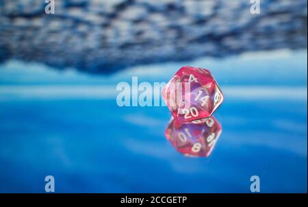 Gros plan d'un dé polyédrique rose transparent sur une surface réfléchissante bleue. Banque D'Images