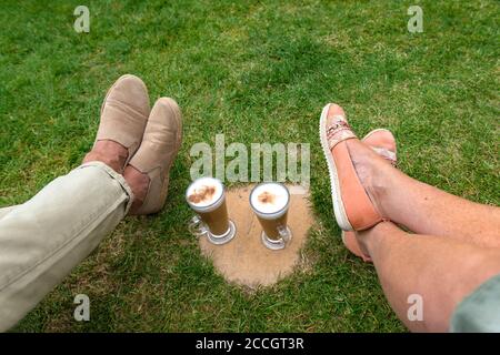 Femme et homme jambes et pieds se détendant dehors sur l'herbe dans le jardin avec 2 délicieux cafés cappuccino dans des tasses en verre sur une dalle de pierre au soleil Banque D'Images