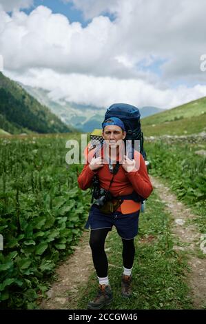 Jeune touriste marchant sur la route de montagne avec un grand sac à dos. Photographe routard voyageant en montagne avec son appareil photo. Banque D'Images