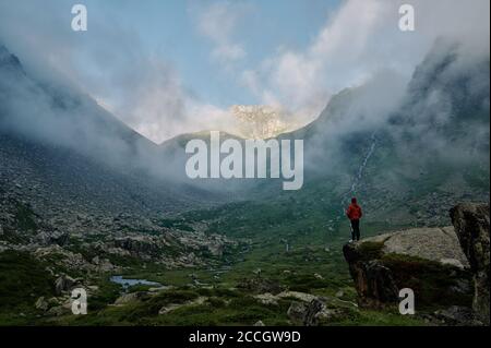 Un jeune homme dans la veste rouge se tient sur le rocher contre la chaîne de montagnes parmi les nuages. Un randonneur extrême dans la nature sauvage. Voyage domestique tre Banque D'Images