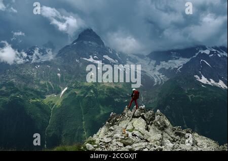 Seul randonneur avec sac à dos et casque contre le paysage sombre de pic de montagne avec le ciel nuageux de tonnerre, les chaînes rocheuses et les pics avec les glaciers. Poste Banque D'Images