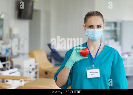 Portrait de la jeune femme médecin en masque et en uniforme tenant la forme du cœur dans ses mains et regardant l'appareil photo Banque D'Images