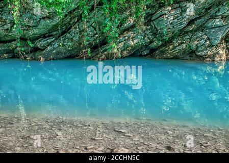 Mouvement rapides flous de la rivière Khosta avec des blocs mouillés et rivage parsemé de feuilles sèches et d'arbres verts ronds Banque D'Images