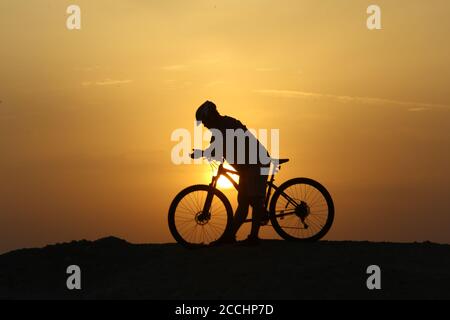 Silhouette d'un jeune homme avec un vélo sur fond d'un coucher de soleil coloré et lumineux. Banque D'Images