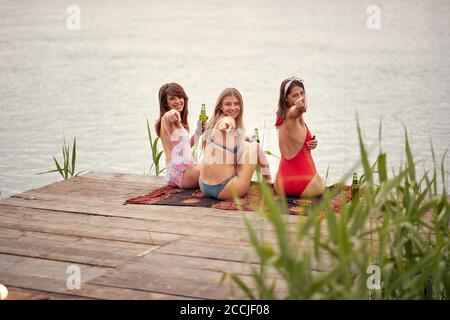 Les jeunes filles séduisantes du bikini se refroidissant avec la bière le bord du lac par temps ensoleillé Banque D'Images