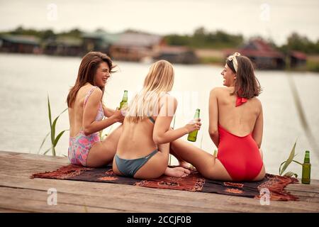 Jeunes filles attrayantes en bikini avec la bière bavardant la rive du lac par temps ensoleillé Banque D'Images