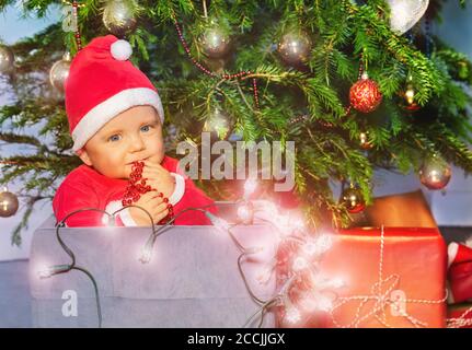 Petit garçon dans un chapeau du Père Noël tenant des lampes illuminées Assis dans une boîte sous l'arbre de Noël Banque D'Images