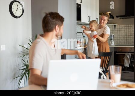 adorable femme et fille parlent avec un homme qui travaille dans la cuisine, un homme s'assoit avec un ordinateur portable. à la maison Banque D'Images