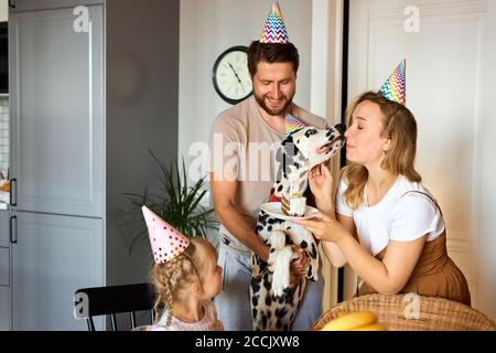 les propriétaires gais et positifs célèbrent l'anniversaire de leur chien, ils félicitent, heureux d'avoir un tel membre de la famille, en chapeaux de fête Banque D'Images
