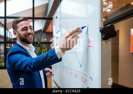 Un homme d'affaires souriant et charmant dessine un graphique sur un tableau blanc pendant la réunion salle du conseil au bureau Banque D'Images