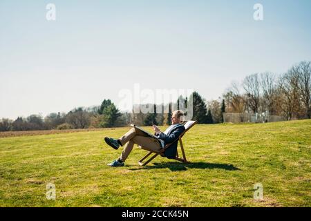 Homme mature professionnel utilisant un smartphone assis sur une chaise avec panneau solaire au parc pendant la journée ensoleillée Banque D'Images