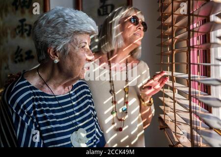 Des amies senior regardent à travers des stores tout en se tenant debout accueil Banque D'Images