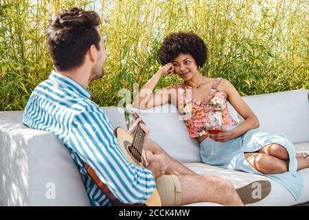 Femme tenant un verre de vin rouge tout en regardant l'homme jouer guitare sur le canapé dans le patio de l'appartement Banque D'Images