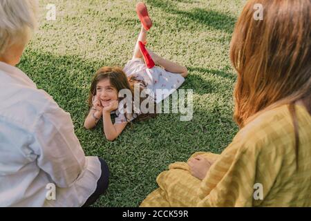 Jolie fille avec les mains sur le menton parlant avec la famille pendant couché sur des terres herbeuses dans la cour Banque D'Images